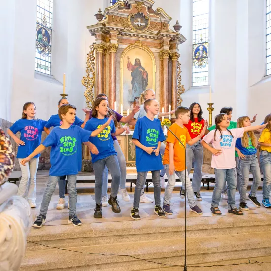 Kinder in bunten T-Shirts stehen singend vor einem Altar in einer Kirche